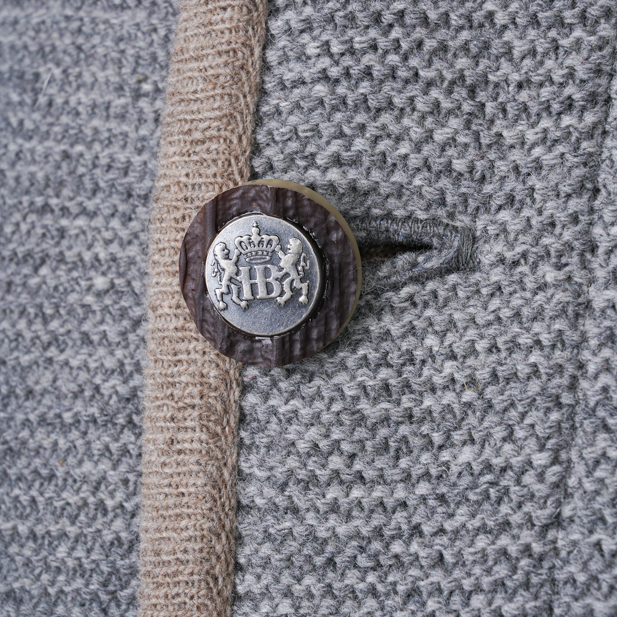 Nahaufnahme eines Knopfes an einer grauen Trachtenjacke mit beige abgesetztem Rand. Der Knopf ist rund und mit einem silberfarbenen Wappenmotiv verziert, auf dem eine Krone und die Buchstaben "HB" zu sehen sind.