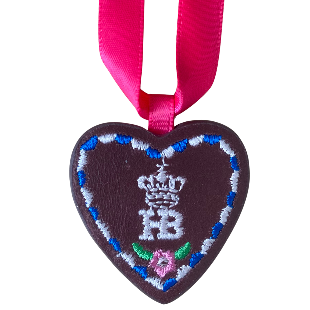 Herzförmiger Anhänger aus braunem Leder mit „HB“-Schriftzug, befestigt an einem pinken Satinband.