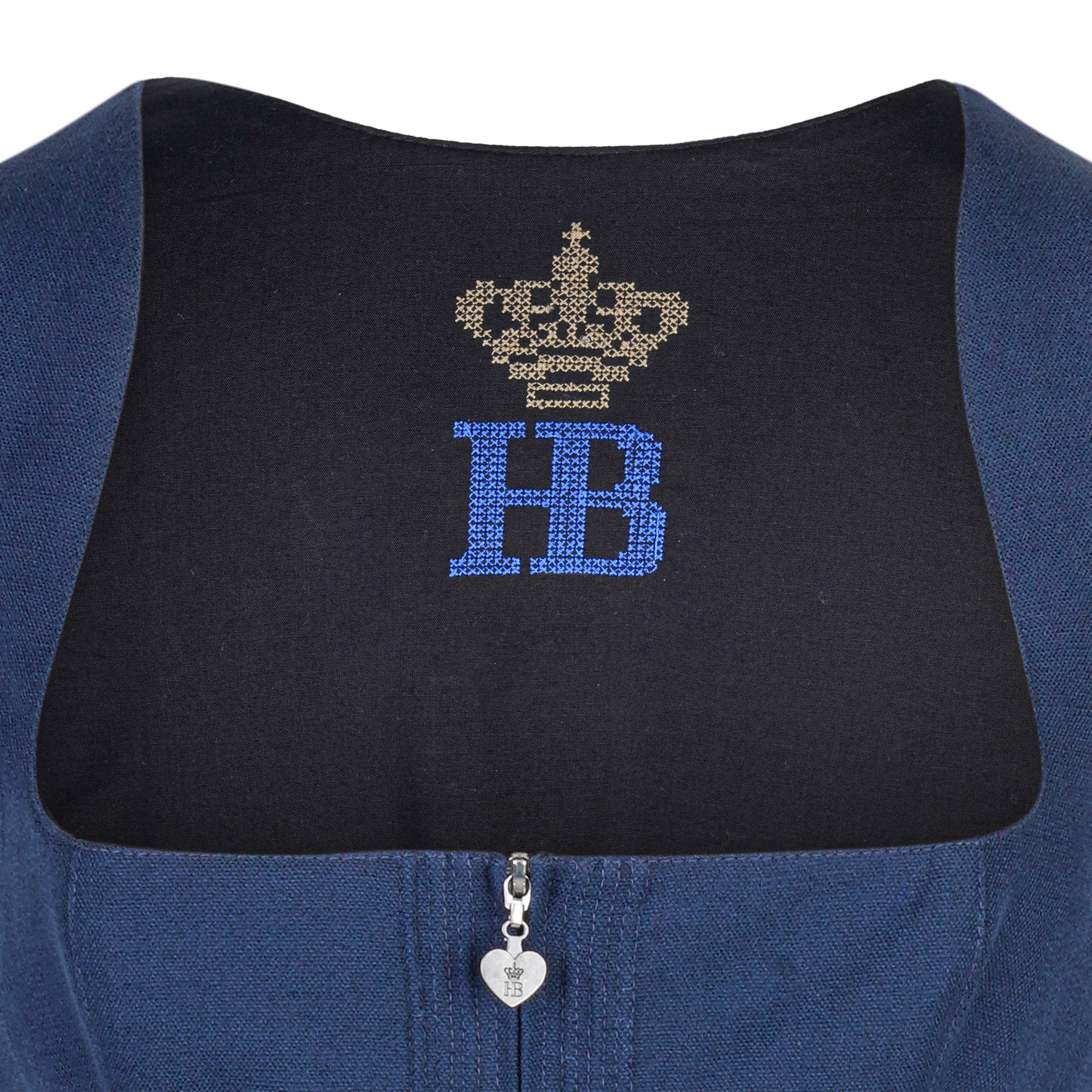 Nahaufnahme des Dirndl-Mieders mit schwarzem Einsatz, darauf ein golden glitzerndes Kronen-Logo und blauer „HB“-Schriftzug aus Strasssteinen; darunter ein silberner Herzanhänger am Reißverschluss.
